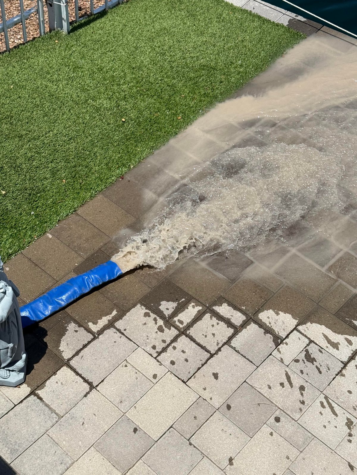 Overhead view of person power washing a patio with concrete pavers, spraying water toward artificial green turf